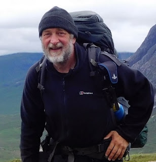 Foreground: A man with a grey and black beard wearing black hiking jacket and hat and carrying a dark blue rucksack on his back. The background is a landscape of hills and valleys